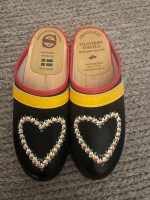 Falsterbo Tofflor med hjärtmotiv - Unika svarta trätofflor från Falsterbo Toffeln med handmålat hjärtmotiv i blommor på ovandelen. Tofflorna har gul och röd kant, samt sula i trä och röd yttersida. Tillverkade i äkta läder och trä, med svensk design och anatomisk fotbädd.