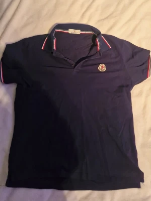 Marinblå pikétröja från Moncler - Snygg marinblå pikétröja från Moncler med klassisk krage och knäppning framtill. Tröjan har röda och vita detaljer på krage och ärmslut samt Moncler-logga på bröstet. Perfekt för en stilren och sportig look.