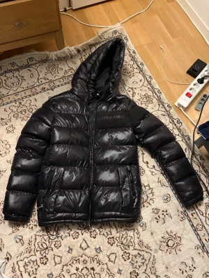Svart pufferjacka från Alessandro - Säljer en svart pufferjacka från Alessandro med glansig finish och stor huva. Jackan har breda, vadderade paneler och dragkedja framtill. Märkeslogga på bröstet och två sidofickor med dragkedja. Perfekt för kalla vinterdagar.