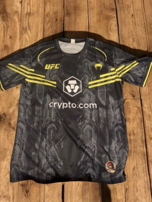 UFC x Venum svart tränings-tshirt - Snygg svart UFC tränings-tshirt från Venum med grått ormmönster och gula detaljer på ärmar och axlar. Stora loggor från UFC och crypto.com framtill. Tillverkad i lätt och snabbtorkande material, perfekt för träning eller gym. KAN KÄNNAS SOM MEDIUM FÖR DEN ÄR COMPRESSED