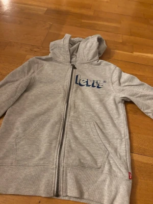 Grå zip hoodie från Levi's - Snygg ljusgrå hoodie från Levi's med dragkedja framtill och blå logga på bröstet. Tröjan har huva och två fickor på sidorna. Perfekt för en avslappnad streetwear-look. Tillverkad i mjukt bomullsmaterial.