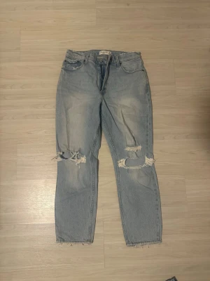 Ljusblå ripped mom jeans från Abercrombie and Fitch - Säljer ett par ljusblå mom jeans med slitningar och stora hål vid knäna. Jeansen har hög midja, fem fickor och råa benslut. Perfekta för en avslappnad och trendig look. Stl 29/32