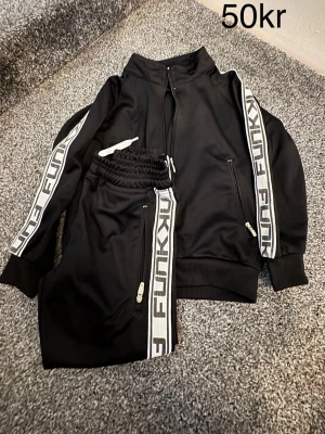 Svart FUNKY tracksuit set - Säljer ett svart tracksuit set från FUNKY med dragkedja på både jacka och byxor. Setet har vita och gråa ränder med texten FUNKY längs ärmar och ben. Perfekt för chill dagar hemma eller när du vill vara bekväm men ändå se cool ut.
