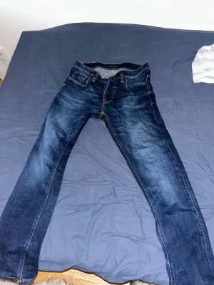 Mörkblå jeans från Nudie Jeans - Säljer ett par mörkblå jeans från Nudie Jeans med klassisk femficksmodell och snygga slitningar. (Grim Tim modellen) Jeansen har raka ben och kontrastsömmar samt Nudie-logga på bakfickan. Perfekta för dig som gillar en avslappnad men stilren look.