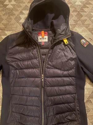 Mörkblå cardigan från Parajumpers - Snygg mörkblå hybridjacka från Parajumpers med huva och quiltad front och rygg. Jackan har dragkedja framtill, bröstficka med dragkedja och en gul detalj vid halsen. Ärmar och sidor är i stretchigt material för extra komfort. Perfekt för blåsiga dagar.