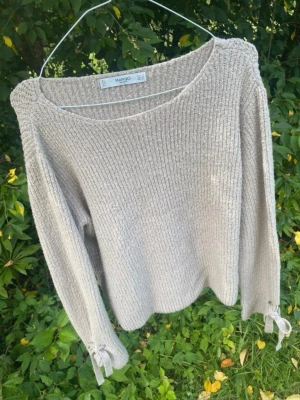 Beige stickad tröja från Mango - Stickad beige tröja från Mango med rund halsringning och långa ärmar. Tröjan har ribbad struktur och dekorativa knytband vid ärmsluten som ger en extra detalj. Perfekt för lager på lager och enkel att matcha med olika stilar.