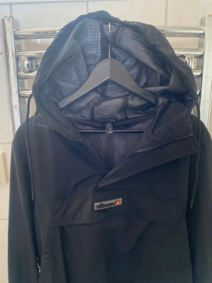 Svart vindjacka från Ellesse - Snygg svart vindjacka från Ellesse med huva och meshfoder. Jackan har dragkedja vid halsen, justerbar huva med snören och Ellesse-logga på bröstet. Perfekt för blåsiga dagar och enkel att matcha med olika outfits.