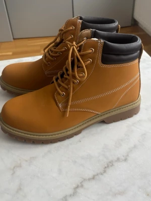 Bruna kängor med snörning - Snygga bruna kängor med snörning och kontrastsömmar. Ovandelen är i mockaliknande material och kragen är vadderad i svart för extra komfort. Grov sula i gummi ger bra grepp. Perfekta för höst och vinter, passar till både jeans och chinos.