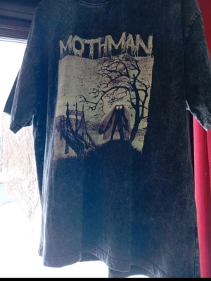 Svart Mothman t-shirt med tryck - Cool svart t-shirt med tvättad look och stort Mothman-tryck framtill. Motivet visar point pleasant bro, träd och den mytomspunna Mothman-figuren. T-shirten har rund hals och korta ärmar, perfekt för dig som gillar mystik och streetwear. I nyskick från Nerdywave Apparel. I storlek Xl. 