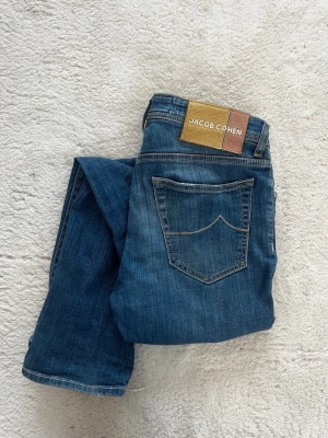 Blå jeans från Jacob Cohen - Snygga blå jeans från Jacob Cohen med klassisk femficksdesign och raka ben. Jeansen har kontrastsömmar och en tydlig patch med logga bak i midjan. Tillverkade i mjukt denimtyg som ger en skön känsla och stilren look.