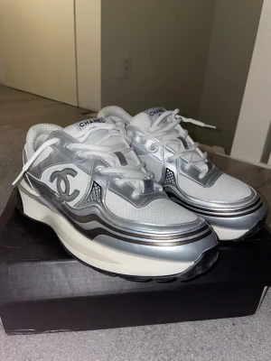 Chanel runners skor - Snygga sneakers från Chanel i vitt och silver med stora loggor på sidan. Skorna har en chunky sula, mesh- och läderdetaljer samt vita skosnören. Perfekta för dig som vill sticka ut med exklusiv design och futuristisk vibe. Pris kan självklart diskuteras!