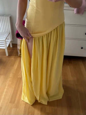 Gul långklänning med snörning - Säljer en elegant gul långklänning med axelbandslös design och asymmetrisk midja. Klänningen har ett matchande scarf.