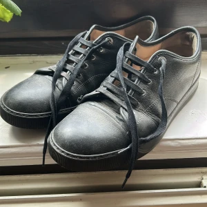 Svarta sneakers från Lanvin - Svarta sneakers i skinn från Lanvin med rund tå och platt sula. Skorna har svarta snören och diskret design utan synliga loggor på utsidan. Passar till många olika stilar och har en klassisk look. Skorna kommer att fräschas upp innan de skickas till kund. Använda en del några defekter men funkar fortfarande bra. Priset kan kanske diskuteras vid lite efterfrågan på skorna. 