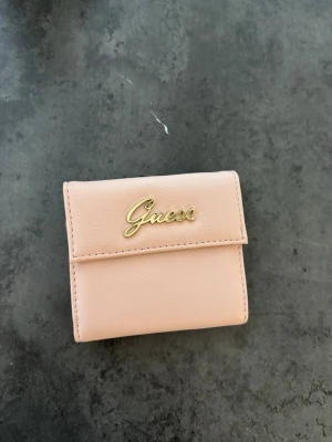 Ljusrosa plånbok från Guess - Snygg ljusrosa plånbok från Guess i mjukt konstläder. Plånboken har guldfärgad logga på framsidan, flera fack för kort och ett myntfack med dragkedja. Smidig och kompakt design som får plats i de flesta väskor. Aldrig använd 