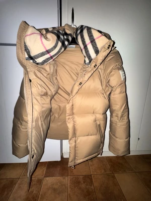 Beige pufferjacka/vest från Burberry - Säljer en beige pufferjacka från Burberry med stor huva och klassiskt rutigt foder. Armana kan tas bort på den och den kan bli vest. Jackan är helt nytt, bara provat på och tagsen finns kvar.