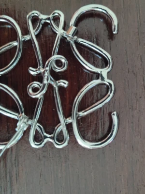 Silverfärgad monogram-brosch - Unik brosch i silverfärgad metall med ett snirkligt monogrammönster. Broschen har en klassisk nål på baksidan och passar perfekt för att ge din outfit en personlig touch. Snygg accessoar för dig som gillar detaljer och stilrena smycken.