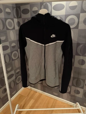 Nike zip hoodie svart/grå - Nike zip hoodie i svart och grått med vit logga på bröstet. Tröjan har hel dragkedja, huva och två sidofickor. Snygg kontrast mellan svart överdel och grå underdel, samt vit detalj på bröstet. Perfekt för en sportig och avslappnad stil.