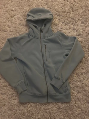 SAILRACING Zip Hoodie - Snygg ljusblå hoodie från Sail racing med hel dragkedja framtill, huva och bröstficka med dragkedja. Tillverkad i mjukt material och har diskret logga på bröstet. Perfekt för en avslappnad och sportig stil.