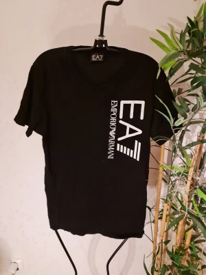 Svart EA7 Emporio Armani t-shirt - Snygg svart t-shirt från EA7 Emporio Armani med stort vitt tryck på framsidan. Klassisk rund halsringning och korta ärmar. Perfekt för dig som gillar stilrena och trendiga plagg med tydlig logga. Tillverkad i mjukt material för skön känsla.