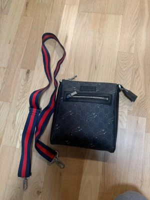 Svart axelväska från Gucci - Snygg svart axelväska från Gucci med klassiskt GG-mönster och silverfärgade detaljer. Väskan har en framficka med dragkedja och en avtagbar axelrem i rött och blått. Tillverkad i canvas och läder, perfekt för dig som gillar exklusiva accessoarer.