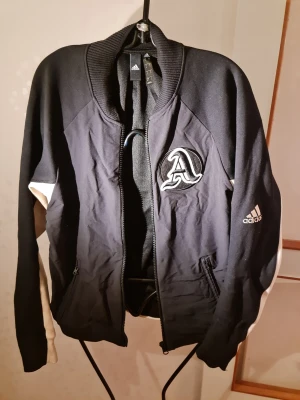 Svart bomberjacka från Adidas - Snygg svart bomberjacka från Adidas med vita detaljer längs ärmarna och ribbad krage. Jackan har dragkedja framtill, två fickor och en stor broderad A-logga på bröstet samt Adidas-logga på ärmen. Perfekt för en sportig och trendig look.