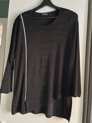 Svart topp med dragkedja från Saphir - Svart topp från Saphir Paris med asymmetrisk nederkant och dekorativa små pärlor framtill. Axelvaddar. Toppen har en unik dragkedjedetalj längs ena axeln och trekvartsärm. Perfekt för dig som gillar en edgy och stilren look. Saknar storleksetikett