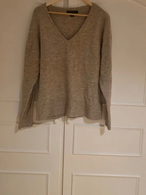 Beige stickad V-ringad tröja - Mysig beige stickad tröja med V-ringning och långa ärmar. Tröjan har ribbstickade muddar vid ärmslut och nederkant. Oversize 