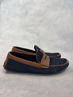 Mörkblå Louis Vuitton loafers med bruna detaljer - Snygga loafers i mörkblått tyg med bruna läderdetaljer runt öppningen och över vristen. Klassisk penny loafer-design med kontrastsömmar och låg profil. Perfekta för en stilren och avslappnad look.