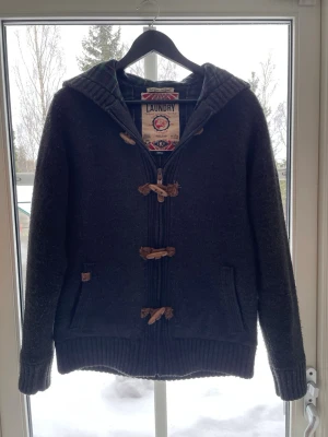 Unik cardigan  - Svart/grå cardigan med träknappar och dragkedja framtill. Tröjan har huva, ribbade muddar och två fickor. Perfekt för kalla dagar och har en avslappnad, stilren look. Nypris -:1599, Strl: S. Vid frågor eller funderingar skriv gärna! Pris inte hugget i sten!!