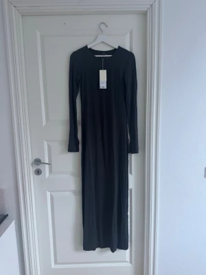 Mörkgrå långärmad maxiklänning - En stilren mörkgrå långklänning med lång ärm och rund halsringning. Klänningen har en smal passform och är tillverkad i ett mjukt, stretchigt material som ger en bekväm känsla. Perfekt för dig som gillar minimalistisk och tidlös stil.