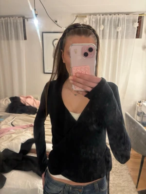 Svart omlottopp med knytband - Snygg svart omlottopp med långa ärmar och knytband i midjan. Toppen har v-ringad halsringning och sitter figurnära. Perfekt att styla med jeans eller kjol för en trendig look. Knyten ser lite konstigt ut men vet inte hur jag ska få den bra, inte använd mycket alls, pris kan diskuteras 
