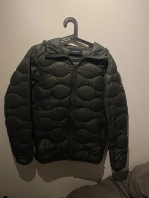 Mörkgrön peak performance dunjacka med huva - Snygg mörkgrön dunjacka från Peak Performance med vågformat quiltat mönster och praktisk huva. Jackan har dragkedja framtill och är lätt vadderad för extra värme. Perfekt för kalla vinterdagar och enkel att matcha med olika outfits.