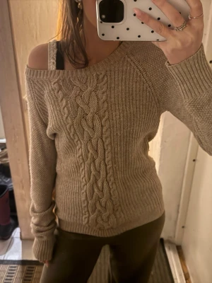 Beige off shoulder stickad tröja - Säljer en beige stickad tröja med breda flätstickade detaljer framtill och ribbade muddar. Tröjan har en avslappnad off shoulder-passform och är perfekt för lager på lager. Mjuk och skön, passar till både jeans och tights.
