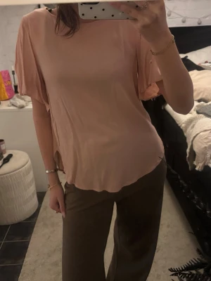 Ljusrosa blus med fladdriga ärmar - Säljer en ljusrosa blus med rund hals och korta, fladdriga ärmar. Blusen har en lös passform och är tillverkad i ett mjukt, tunt material som ger ett snyggt fall. Perfekt att styla med både jeans och kjol.