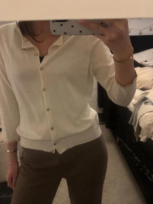 Krämvit kofta med knappar - Stilren krämvit kofta med rund halsringning och knäppning framtill. Koftan har trekvartsärmar och ribbade muddar vid ärmslut och nederkant. Perfekt att matcha med både jeans och kjol för en clean look.