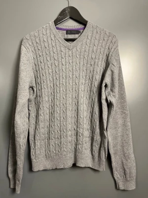 Grå kabelstickad tröja Brothers - Snygg grå kabelstickad tröja från Brothers i 100% bomull. Tröjan har V-ringning, långa ärmar och ribbade muddar vid ärmslut och nederkant. Perfekt för lager på lager och enkel att matcha med jeans eller chinos.