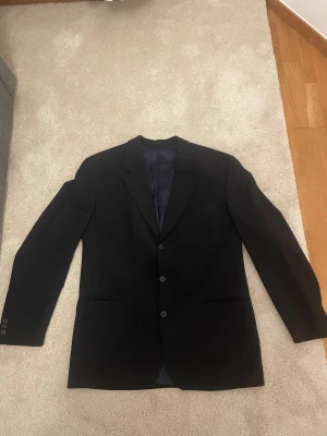 Svart kavaj från Oscar Jacobson - Klassisk svart kavaj från Oscar Jacobson i Super 100's ull. Kavajen har tre knappar framtill, två fickor och en stilren passform. Perfekt för en dressad look och tillverkad i 100% ren ull för en lyxig känsla.