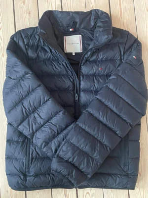 Svart pufferjacka Tommy Hilfiger - Snygg svart pufferjacka från Tommy Hilfiger med diskreta loggor på bröstet och ärmen. Jackan har hög krage, dragkedja framtill och två sidofickor. Perfekt för kalla dagar med sin vadderade design och klassiska look.