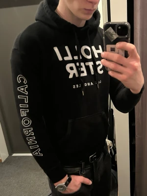 Svart Hollister hoodie med tryck - Svart hoodie från Hollister med stor vit logga på bröstet och texten 'CALIFORNIA' längs ena ärmen. Klassisk känguruficka framtill och justerbar huva med snören. Perfekt för en avslappnad och cool stil.