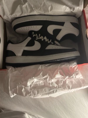 Nike Dunk Low svart/vit sneaker - Snygga Nike Dunk Low sneakers i svart och vitt läder med klassisk swoosh på sidan. Skorna har vita skosnören och en vit sula med svart yttersula. Perfekt för dig som gillar stilrena och tidlösa sneakers med låg profil.