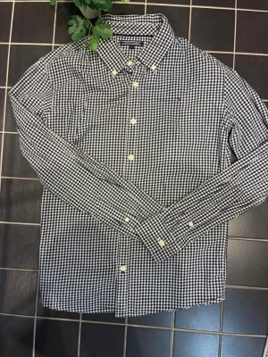 Rutig skjorta från Tommy Hilfiger - Klassisk svartvit rutig skjorta från Tommy Hilfiger med button-down krage och vita knappar. Skjortan har lång ärm och liten broderad logga på bröstet. Tillverkad i mjuk bomull som känns skön mot huden. Perfekt för en clean och stilren look.