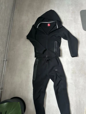 Svart Nike tracksuit  - Säljer en svart zip hoodie från Nike med huva och dragkedja framtill. Hoodien har diskret Nike-logga på bröstet och praktiska fickor med dragkedja. Tillverkad i mjukt och bekvämt material, perfekt för en avslappnad look