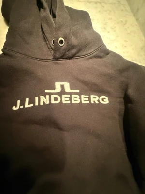 Svart hoodie från J.Lindeberg - Snygg svart hoodie från J.Lindeberg med stor logga tryckt på bröstet. Klassisk modell med huva och dragsko, samt metallöljetter. Mjuk insida och ribbade muddar. Perfekt för en avslappnad och stilren look.