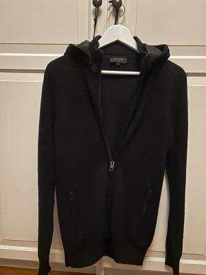 Svart cardigan från inbicode  - Säljer svart cardigan med dragkedja framtill och två fickor . Tröjan har en huva med glansig detalj och ribbade muddar vid ärmslut och nederkant. Perfekt för dig som gillar en enkel och stilren look.