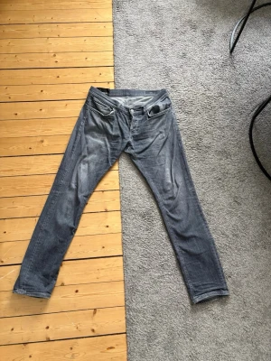 Gråa dondup jeans - Säljer ett par gråa jeans från dundup i modellen George och storlek 34. Jenssen är använda men är fortfarande i gott skick det ända är att dondup loggan som ska sitta på bakfickan har trillat av men om man kan hitta en sådan så är det enkelt att sätta på den igen. Dem är stretchiga 