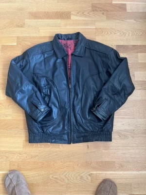 Svart skinnjacka från wild cats - Snygg svart skinnjacka med dragkedja framtill och två stora fickor. Insidan har ett unikt rött foder med tryck i svart. Jackan har klassisk krage, justerbara ärmslut och resår i midjan för en schysst passform. Perfekt för dig som vill ha en tidlös och cool look.