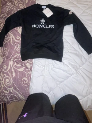 Svart Moncler sweatshirt - Snygg svart sweatshirt från Moncler med vit logga och text framtill. Klassisk rund halsringning och ribbade muddar vid ärmslut och nederkant. Perfekt för en stilren och trendig look.