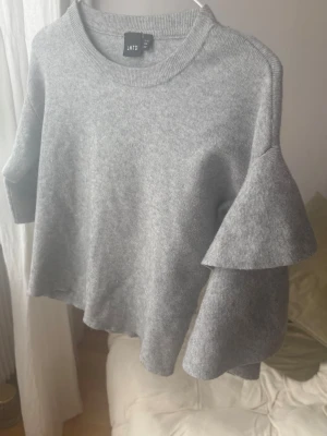 Grå stickad tröja med volangärm LMTD - Snygg grå stickad tröja från LMTD med trendiga volangärmar och rund halsringning. Tröjan har en rak, lite kortare passform och är perfekt för dig som gillar en stilren men ändå unik look.