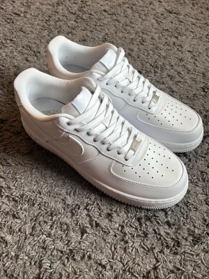 Nike Air Force 1 vita sneakers - Klassiska Nike Air Force 1 sneakers i helvitt läder med perforerad tåbox, vit sula och vita skosnören. Ikonisk siluett med låg profil och diskret Nike-logga på sidan. Perfekta för dig som gillar en clean och tidlös stil. Finns även box till och creas skydd