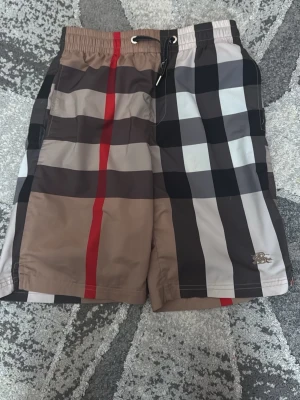 Rutig badshorts från Burberry - Säljer ett par klassiska badshorts från Burberry med ikoniskt rutigt mönster i beige, svart, vitt och rött. Shortsen har elastisk midja med snörning och sidoficka. Materialet är lätt och snabbtorkande, perfekt för stranden eller poolen. Burberry-logga broderad på benet.
